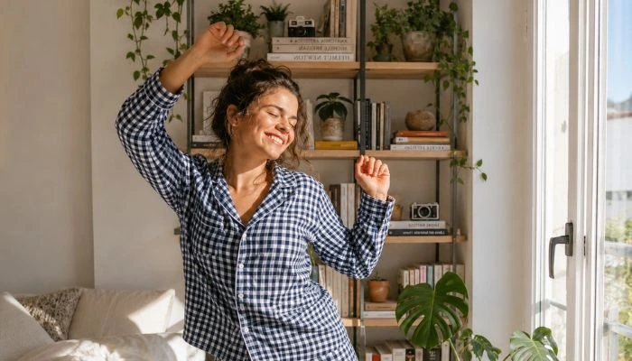 Una mujer feliz baila en pijama de cuadros dentro de una habitación luminosa con libros y plantas - Opiniones Algodonea